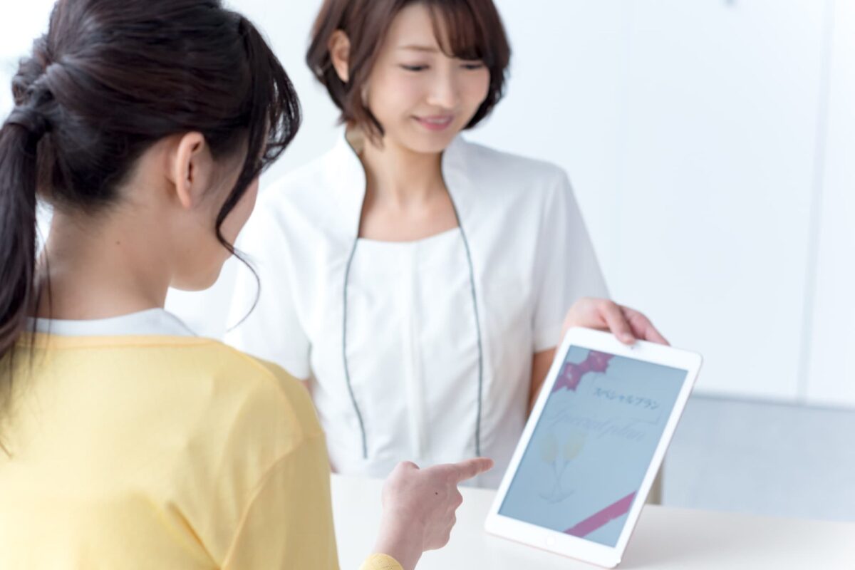 白いユニフォームのスタッフがタブレットで施術メニューを説明するカウンセリング風景